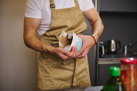 Close-up man chef in beige apron, pouring some salt from paper package, seasoning meal while cooking...