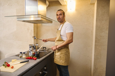 Caucasian handsome young chef man 40s, dressed in beige apron, standing at kitchen counter, cooking delicious dinner for his family, smiling and confidently looking at camera. Ingredients on the tableの写真素材