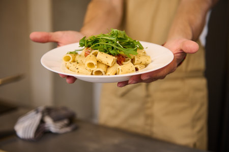 Selective focus on male chef hands holding a white plate with Italian pasta with tomato sauce, seasoned with basil and arugula leaves on chef's hands. Italian cuisine. Culinary. Food backgroundの写真素材
