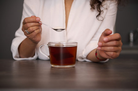 Close-up woman's hands holding a teaspoon with white sugar above a glass cup of freshly made black tea in the home kitchen, preparing hot drink for breakfast in the morningの写真素材