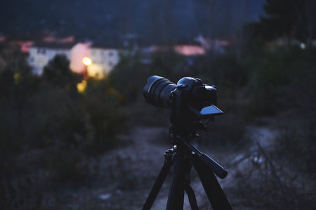 A digital camera with black mockup touch screen placed on tripod by a travel photographer hiker, capturing the beautiful view of a city in mountains at sunrise. The concept of World photography Dayの写真素材