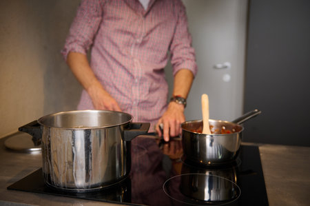 Focus on steel saucepan on the induction oven, electric stove against blurred male chef switching on panel for boiling water. Cropped view of blurred man switching on, turning on the electric stoveの写真素材