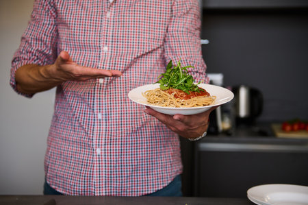 Close-up hands of young Italian chef showing a white plate with Italian spaghetti capellini, standing in the home kitchen. Chef showing served dish with Italian pasta. Italy. Food. Culture. Traditionsの写真素材