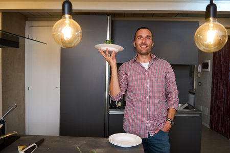 Authentic portrait of a young male chef holding a plate with Italian pasta, smiling looking at camera, standing in the minimalist home kitchen interior. People. Italian culture. Lifestyle. Culinaryの写真素材
