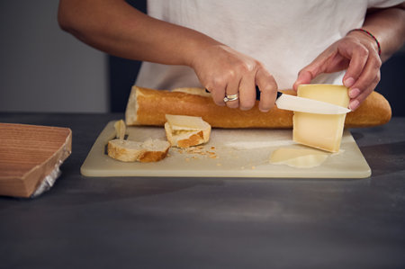 Close-up hands of a woman in white t-shirt, cutting cheese and bread, preparing sandwiches for snack, standing at kitchen countertop with fresh organic ingredients. Food and drink conceptの写真素材