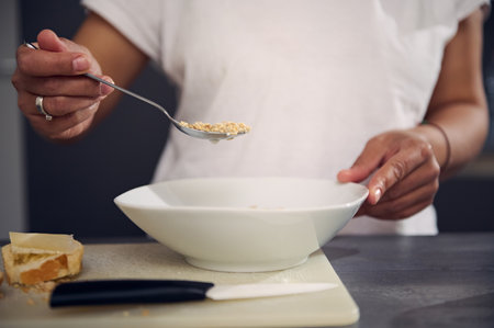 Details on the hand of woman in white t-shirt, holding a spoon with wholegrain muesli granola above a white bowl, enjoying a healthy breakfast in the morning. Healthy eating, slimming and diet conceptの写真素材
