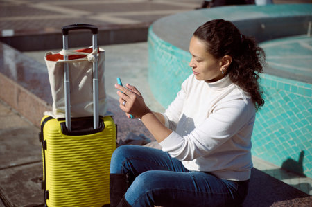 Woman sitting next to a yellow suitcase by a fountain, using a smartphone. The image conveys travel, relaxation, and connectivity in an outdoor urban environment on a sunny day.の写真素材