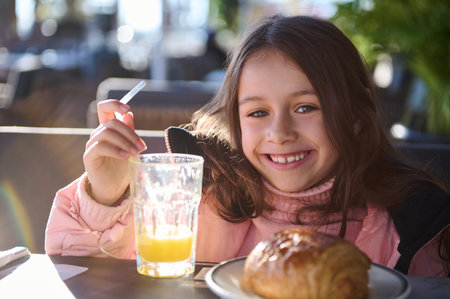Happy young girl enjoying a sunny morning breakfast at an outdoor cafe. She is smiling brightly,...