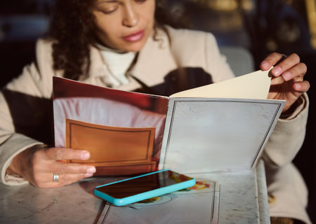 A woman sits at an outdoor cafe, reading a menu with a focused expression. A smartphone rests on the table beside her. The afternoon sun casts a warm glow on the scene.の写真素材
