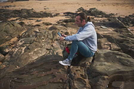 A man in casual clothes sits on a rocky beach and uses a tablet. He appears focused, perhaps working or enjoying casual browsing while surrounded by the beauty of nature.の写真素材