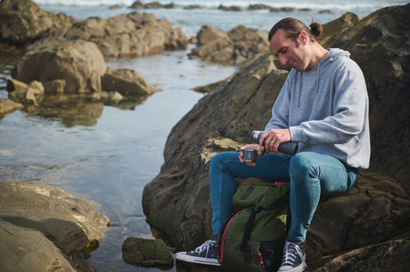 A man in casual attire sits on rocks near the sea, pouring a hot drink from a thermos into a cup. The setting is serene and suggests a peaceful outdoor adventure.の写真素材