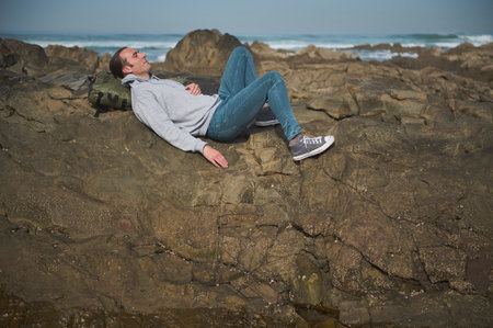A man is lying on a rugged rocky beach, relaxing and gazing at the serene ocean waves. He is wearing casual clothing and appears calm and content, embracing the tranquility of nature around him.の写真素材