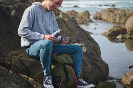 A man in casual attire sits on a rocky coast, taking a break and enjoying a hot drink from a thermos bottle. He has a backpack beside him and the ocean in the background.の写真素材