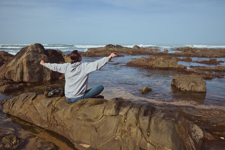 A person in a gray hoodie stretches while sitting on a rock by the sea. The scene features a clear blue sky, rocky coastline, and the calm, serene ocean waves creating a peaceful atmosphere.の写真素材