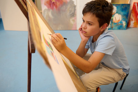 Kids have art class, drawing, painting with teacher in workshop. Young boy is deeply focused on his artwork, showing creativity and concentrationの写真素材