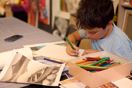 Kids have art class, drawing, painting with teacher in workshop. A young boy focuses on his drawing with a variety of colorful pencils in front of him on a cluttered table.の写真素材