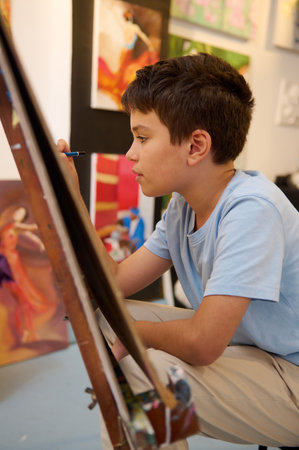 A young boy attentively painting on a canvas during an art class. He is deeply engrossed in his creative process, surrounded by colorful artworks in the background.の写真素材