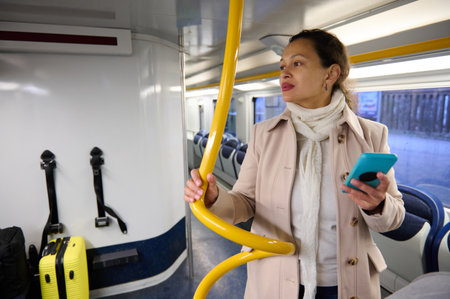 A woman holds a smartphone and stands inside a train next to her luggage. She appears focused and is dressed warmly in a coat and scarf.の写真素材