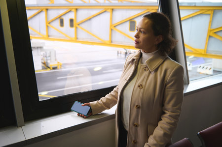 Woman in beige coat holding a smartphone, gazing thoughtfully out of an airport terminal window towards the runway and parked aircraft.の写真素材