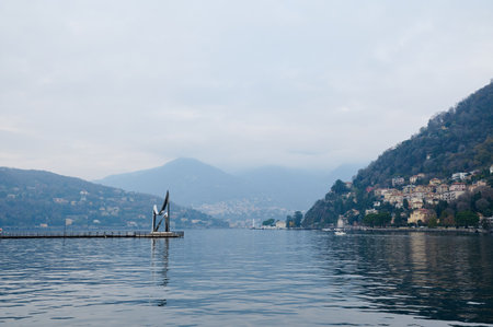 Scenic panorama of Lake Como with tranquil waters, atmospheric mist over the mountains, and a picturesque lakeside town. Ideal for travel and nature enthusiasts.の写真素材