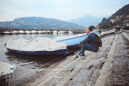 A man sits by the lakeside with docked boats, gazing at the surrounding mountains and reflecting on the peaceful atmosphere. Perfect for themes of travel, relaxation, and contemplation.の写真素材