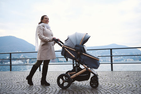 A woman in a light coat and scarf walks with a baby stroller along a lakeside path. Serene landscape...