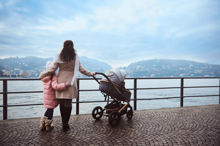 A mother walks with her child and baby stroller by a scenic waterfront on a cloudy day, enjoying...