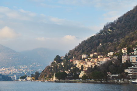 Beautiful village by a lakeside surrounded by steep, forested hills. The calm water reflects the serene atmosphere under a cloudy sky.の写真素材
