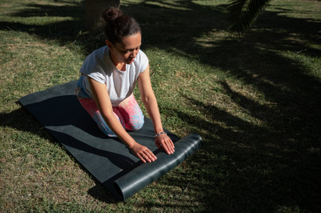 A woman rolling a yoga mat in a park on a sunny day, preparing for an outdoor exercise session. Calm and focused moment.の写真素材