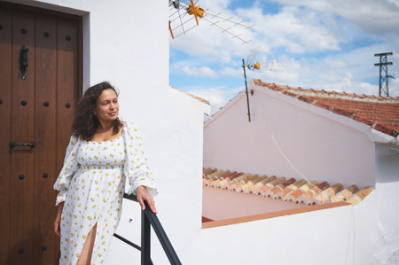 Woman in a white dress with floral pattern standing outside a traditional Mediterranean house. Wooden door and blue sky create an inviting atmosphere.の写真素材