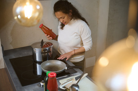 Woman cooking in a modern kitchen, holding a bottle of sauce and checking cookware on an electric stove. Warm lighting adds ambiance.の写真素材