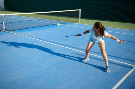 A young woman in motion hitting a tennis ball during a game on an outdoor blue tennis court, displaying agility and determination.の写真素材
