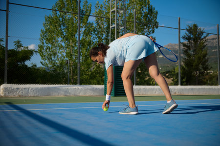 A middle-aged woman in her 40s is playing tennis on an outdoor court. The image captures her in action, bending down to pick up a tennis ball, reflecting her athleticism and love for sports.の写真素材