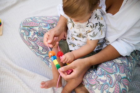 Top view of a mother hugging her baby while he plays with educational wooden toys. A cute boy touches a chain with wooden pendants and studies new objects by touch. Motor skills development conceptの写真素材