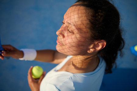 Close-up of a woman holding a tennis ball, with a racket casting shadows on her face on a sunny day. A moment of focus and concentration before serving.の写真素材