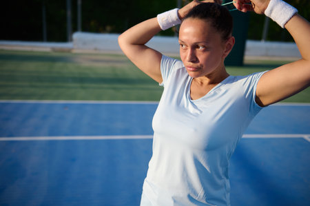 Focused female tennis player tying her hair on a blue court while getting ready for a game under the...