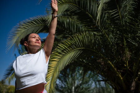 A woman stretches her arm up while practicing yoga outdoors under a palm tree, embracing a serene and mindful moment in nature on a bright, sunny day.の写真素材