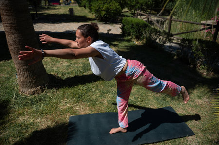 A woman holding a yoga pose on a mat outdoors in a park. She is focused and balanced, enjoying the calm and peaceful environment while engaging in fitness and wellness activities.の写真素材