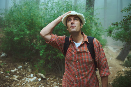 Male hiker in a misty forest, wearing a hat and plaid shirt, looking up thoughtfully. Outdoor adventure and nature exploration.の写真素材
