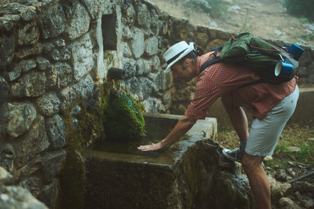 A hiker in nature wearing a hat and backpack, stopping to drink water from a stone fountain. The scene is surrounded by moss and rustic stone walls, symbolizing exploration and outdoor adventure.の写真素材