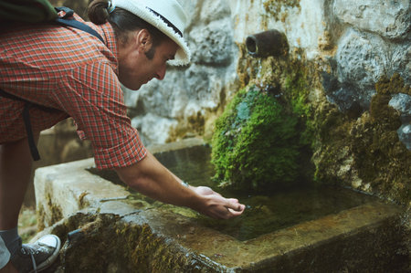 A hiker in a white hat and plaid shirt bends down to drink fresh water from a stone spring surrounded by moss. Nature, refreshment, and adventure.の写真素材