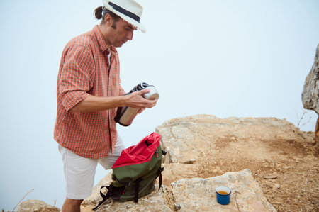 A man in casual attire and a hat pouring a drink from a thermos while taking a break on a rocky mountain top during a hiking trip. A cup and a backpack are seen nearby.の写真素材