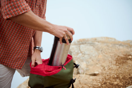 Hiker packing a stainless steel thermos into a green backpack on a rocky trail. Outdoor adventure, travel, and exploration concept.の写真素材