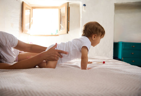 Parent helping baby learn to crawl on a cozy bed in a sunlit bedroom. Baby with supportive hands, open window in background.の写真素材