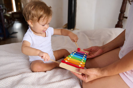 Adorable baby playing a colorful xylophone alongside a parent. Capturing early childhood development and music education moments at home in a cozy bedroom setting.の写真素材