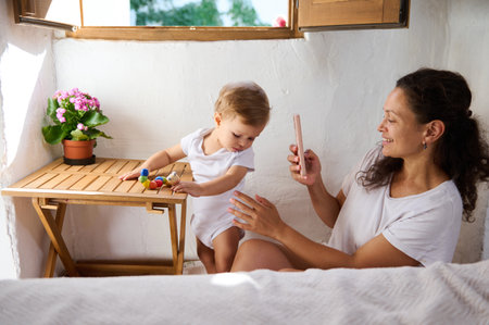Happy mother taking a picture of her adorable baby boy playing with colorful toys on a wooden table....