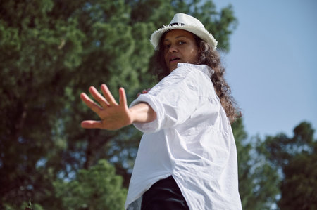 A confident woman in a white shirt and hat stands outdoors, raising her hand as if to stop, with a...