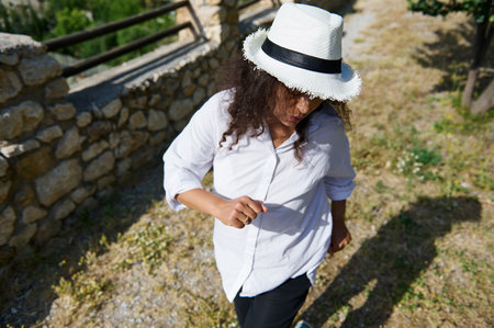 A person with long hair, wearing a white shirt and hat, walks outdoors along a stone wall on a sunny day, enjoying the summer weather.の写真素材
