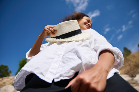 Low angle view of a woman holding a white hat and looking happy against a clear blue sky. Perfect for depicting outdoor fun, sunny weather, and carefree lifestyle.の写真素材