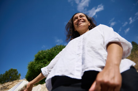 A joyful woman smiles brightly while enjoying a sunny day outdoors. Shot from a low angle against a...